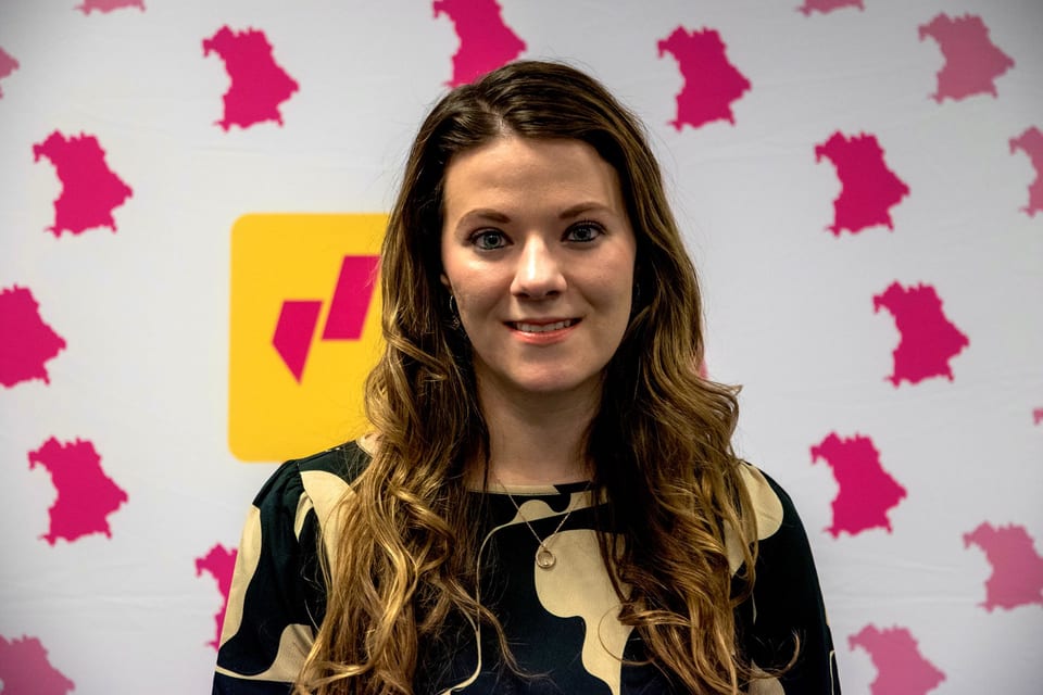 Porträt von Angela Rehekampff mit langen, gewellten Haaren vor einer weißen Fotowand mit pinkfarbenen Umrissen Bayerns und dem gelben Logo der FDP Bayern